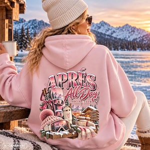 Könnte beinhalten: Ein hellrosa Kapuzenpullover mit einem Grafikdruck auf der Rückseite und dem Text APRÉS All Day SOCIAL CLUB. Das Motiv zeigt ein Snowboard, Skistöcke, eine Mütze, eine Skibrille, ein Getränk und ein Holzschild vor einer verschneiten Bergkulisse.