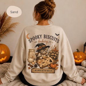 Könnte beinhalten: Ein sandfarbenes Sweatshirt mit Rundhalsausschnitt und einem Grafikdruck auf der Rückseite. Das Motiv zeigt ein Kätzchen mit Hexenhut, das Kekse mit Mehl und Kürbissen backt. Der Text lautet: SPOOKY BISCUITS bakery und WE KNEAD EM YOU NEED EM.