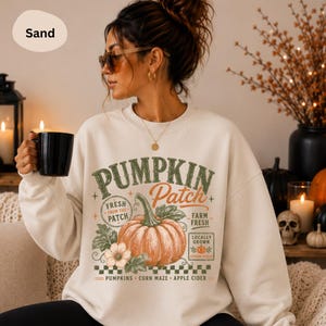 Könnte beinhalten: Ein sandfarbenes Sweatshirt mit Rundhalsausschnitt und einem Grafikdruck mit dem Text PUMPKIN Patch, FRESH FROM THE PATCH, FARM FRESH, LOCALLY GROWN, PUMPKINS, CORN MAZE und APPLE CIDER, zentriert um eine große orangefarbene Kürbisillustration.