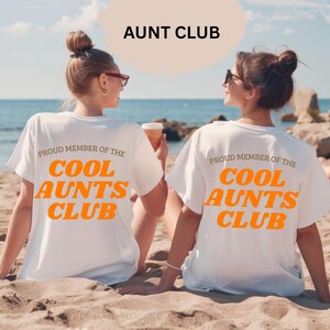 May include: Two women on a beach wearing white t-shirts. The shirts display the text "PROUD MEMBER OF THE COOL AUNTS CLUB" in orange. Above them is the text "AUNT CLUB". The background shows the sea and sand.