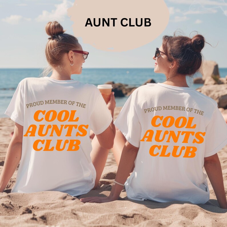 May include: Two women on a beach wearing white t-shirts. The shirts display the text "PROUD MEMBER OF THE COOL AUNTS CLUB" in orange. Above them is the text "AUNT CLUB". The background shows the sea and sand.