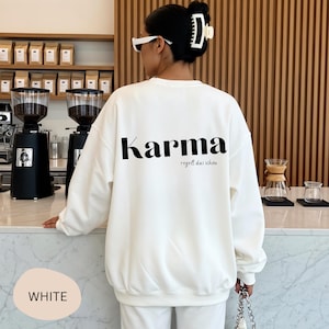 Könnte beinhalten: Weißes Sweatshirt mit dem Wort "Karma" in Schwarz auf dem Rücken aufgedruckt, zusammen mit dem Satz "regelt das schon". Das Sweatshirt ist Teil eines weißen Outfits. Das Bild zeigt auch eine Kaffeehaus-Umgebung.