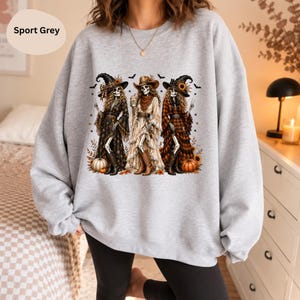 Könnte beinhalten: Ein sportgraues Sweatshirt mit Rundhalsausschnitt und einem Grafikdruck von drei Skelettfiguren in Westernkleidung, einschließlich Hüten und Stiefeln, umgeben von Kürbissen, Sonnenblumen und Fledermäusen. Der Text Sport Grey ist sichtbar.