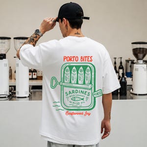Könnte beinhalten: Ein weißes kurzärmeliges T-Shirt mit einem großen Aufdruck auf der Rückseite. Das Motiv zeigt eine grüne Sardinendose mit dem Text PORTO BITES, SARDINES WILD CAUGHT IN OLIVE OIL, PORTO und Seaflavored Jay in roter und grüner Farbe.