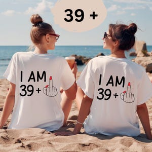 Könnte beinhalten: Zwei weiße T-Shirts mit dem Text "I AM 39+" und einer Mittelfinger-Grafik. Die Shirts werden von zwei Personen am Strand getragen. Die Zahl "39+" steht ebenfalls über den Personen.