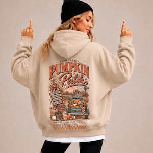 Könnte beinhalten: Ein beigefarbener Kapuzenpullover mit einem Grafikdruck auf der Rückseite und dem Text "FARM FRESH PUMPKIN PATCH". Das Motiv zeigt einen mit Kürbissen beladenen Lastwagen, eine Scheune, Maispflanzen und Schilder mit der Aufschrift "PUMPKINS", "CORN MAZE", "APPLE CIDER", "HAYRIDES".