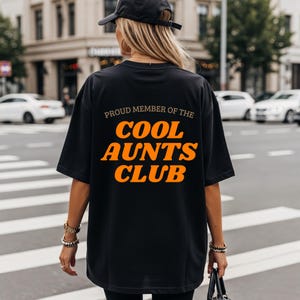 May include: Black t-shirt featuring the orange text "PROUD MEMBER OF THE COOL AUNTS CLUB". The shirt is a relaxed fit, ideal for casual wear. The image shows the back of the shirt.