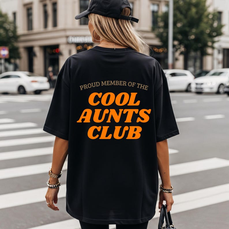 May include: Black t-shirt featuring the orange text "PROUD MEMBER OF THE COOL AUNTS CLUB". The shirt is a relaxed fit, ideal for casual wear. The image shows the back of the shirt.