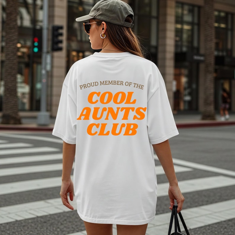 May include: White t-shirt featuring the text "PROUD MEMBER OF THE COOL AUNTS CLUB" in orange and grey lettering. The shirt has a relaxed, oversized fit.