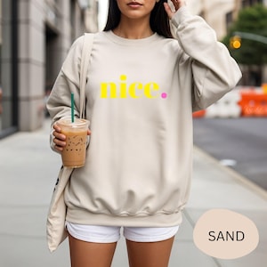 Könnte beinhalten: Ein sandfarbener Pullover mit dem Wort "nice." in Gelb und Pink aufgedruckt. Der Pullover hat lange Ärmel und einen Rundhalsausschnitt. Das Model trägt weiße Shorts und hält einen transparenten Plastikbecher mit einem grünen Strohhalm.