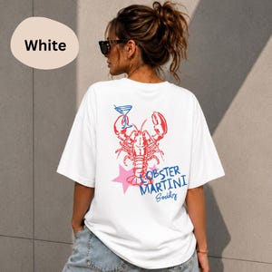 Op de afbeelding: Een wit t-shirt met een rode kreeftafbeelding die een blauw martiniglas vasthoudt. Onder de kreeft staat een roze ster en de blauwe tekst "LOBSTER MARTINI Society". Het ontwerp is gecentreerd op de achterkant van het t-shirt.
