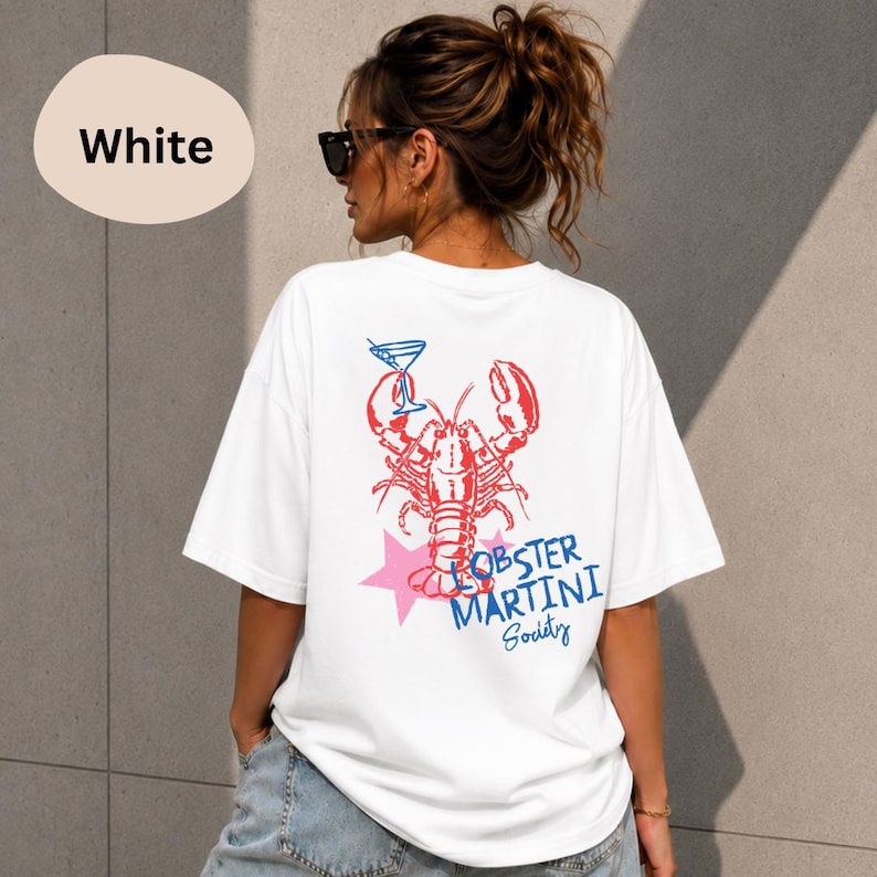 Op de afbeelding: Een wit t-shirt met een rode kreeftafbeelding die een blauw martiniglas vasthoudt. Onder de kreeft staat een roze ster en de blauwe tekst "LOBSTER MARTINI Society". Het ontwerp is gecentreerd op de achterkant van het t-shirt.