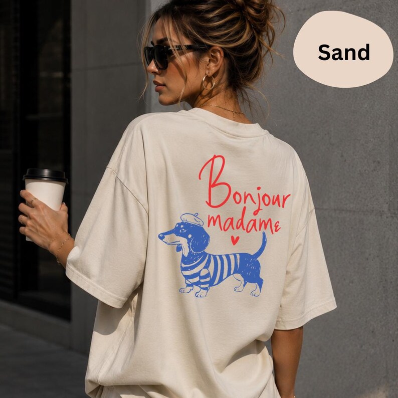 May include: A sand-coloured t-shirt featuring a graphic on the back with a blue dachshund wearing a beret and a striped shirt. The text above the dog reads Bonjour madame in red script, accompanied by a small red heart icon below the text.