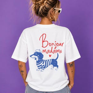 May include: A white short-sleeved t-shirt featuring a graphic of a dachshund wearing a blue and white striped shirt and a beret. Above the dog, the text reads Bonjour madame in red script, accompanied by a small red heart icon below the text.
