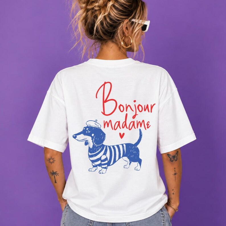 May include: A white short-sleeved t-shirt featuring a graphic of a dachshund wearing a blue and white striped shirt and a beret. Above the dog, the text reads Bonjour madame in red script, accompanied by a small red heart icon below the text.