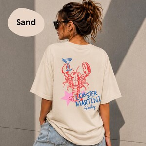 Op de afbeelding: Een zandkleurig t-shirt met een afbeelding op de achterkant van een rode kreeft, een blauw martiniglas, een roze ster en de blauwe tekst "LOBSTER MARTINI Society". Het t-shirt heeft korte mouwen en een ronde hals, gezien vanaf de achterkant.