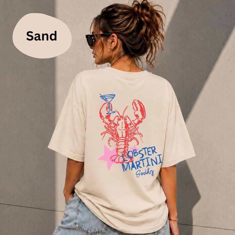 Op de afbeelding: Een zandkleurig t-shirt met een afbeelding op de achterkant van een rode kreeft, een blauw martiniglas, een roze ster en de blauwe tekst "LOBSTER MARTINI Society". Het t-shirt heeft korte mouwen en een ronde hals, gezien vanaf de achterkant.