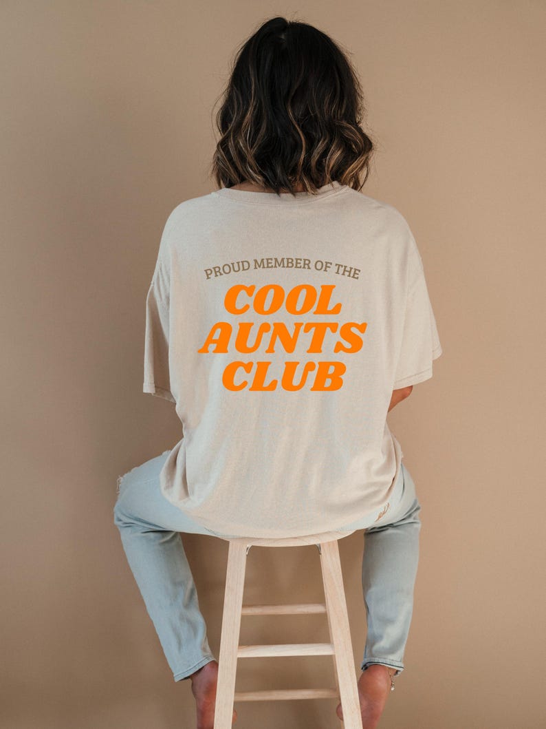 May include: A beige t-shirt with the orange text "PROUD MEMBER OF THE COOL AUNTS CLUB". The person is wearing light blue jeans and is sitting on a wooden stool. The background is a plain tan colour.