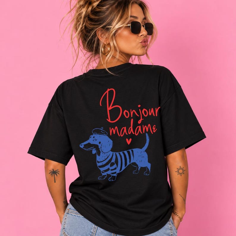 May include: A black short-sleeved t-shirt featuring a graphic of a blue dachshund wearing a beret. The text "Bonjour madame" is printed in red above the dog, accompanied by a small red heart. The shirt has a crew neckline and a loose fit.