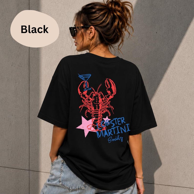Op de afbeelding: Een zwart t-shirt met korte mouwen en een afbeelding op de achterkant. Het ontwerp toont een rode kreeft die een blauw martiniglas vasthoudt, een roze ster en de blauwe tekst "LOBSTER MARTINI Society". Het shirt heeft een ronde hals en een wijde pasvorm.