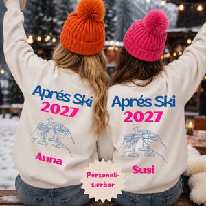 Könnte beinhalten: Zwei cremefarbene Sweatshirts mit dem blauen Schriftzug "Aprés Ski" und dem pinken "2027" über einer blauen Strichzeichnung von anstoßenden Cocktailgläsern. Die Namen "Anna" und "Susi" sind darunter in Pink gedruckt. Dazu wird jeweils eine Bommelmütze getragen.