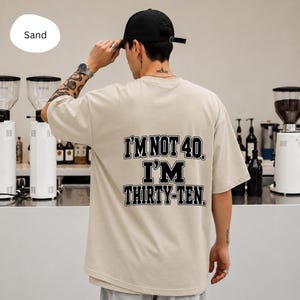 Peut inclure: Un t-shirt à manches courtes de couleur sable avec un texte noir de style universitaire au dos qui indique, I'M NOT 40. I'M THIRTY-TEN. Le t-shirt présente une coupe ample et décontractée, fabriqué dans un tissu uni avec un col rond simple.