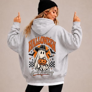 Könnte beinhalten: Ein hellgrauer Kapuzenpullover mit einem Rückendruck, der ein Gespenst mit Cowboyhut zeigt, das einen Kürbis hält. Der Text lautet HALLOWEEN und Spooky Vibes Only, umgeben von Fledermäusen, Sternen und schwarz-orangen Karomustern.