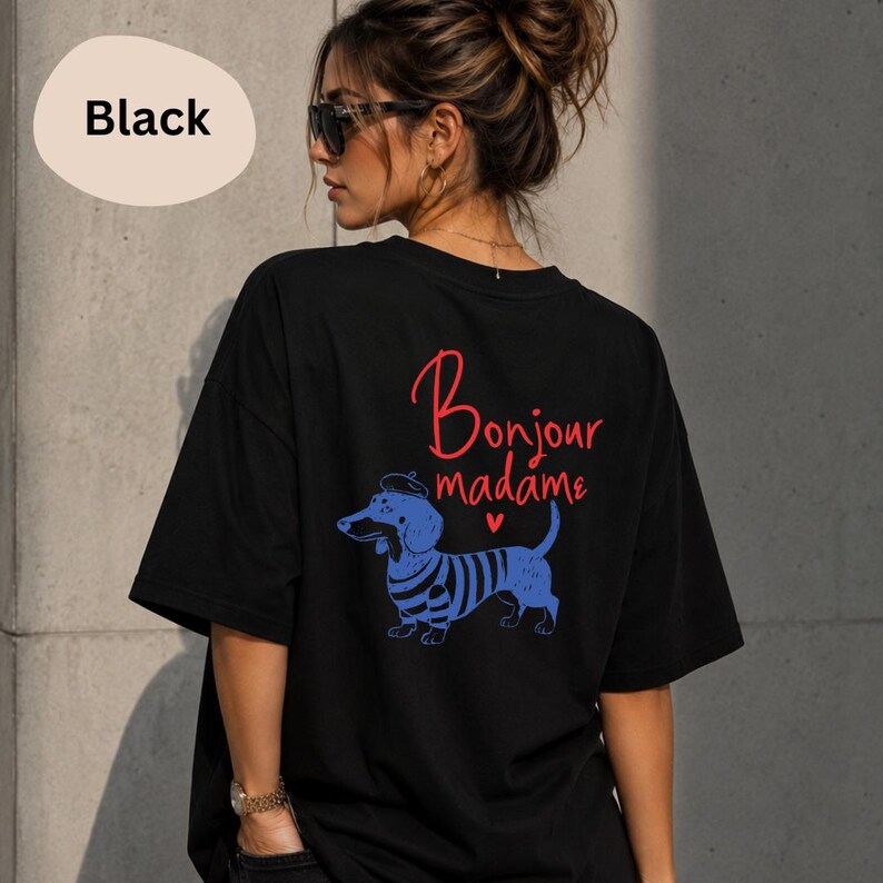 May include: A black short-sleeved t-shirt featuring a graphic on the back. The design includes the red text Bonjour madame above a blue illustration of a dachshund wearing a beret and a striped shirt, accompanied by a small red heart symbol.