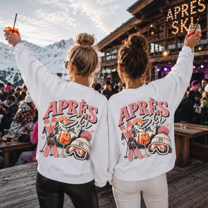Pode incluir: Duas camisolas brancas de gola redonda com uma impressão gráfica nas costas. O design inclui o texto "APRÈS Ski" em rosa, esquis cor-de-rosa cruzados, um copo de cocktail e um gorro de inverno com óculos, sobre uma paisagem de montanha.