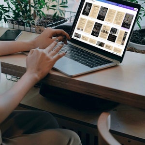 K&ouml;nnte beinhalten: Eine Person arbeitet an einem silbernen Laptop mit einem wei&szlig;en Bildschirm, auf dem ein digitales Dokument mit mehreren Seiten angezeigt wird. Das Dokument hat einen braunen Hintergrund mit schwarzem Text und Bildern. Der Laptop steht auf einem hellbraunen Holztisch.