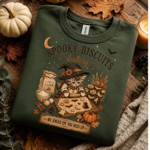 Könnte beinhalten: Ein dunkelolivgrüner Rundhals-Pullover mit einer Grafik eines Kätzchens mit Hexenhut, das Teig mit Kürbis- und Fledermausformen ausrollt. Der Text lautet SPOOKY BISCUITS bakery und WE KNEAD EM YOU NEED EM in einer rustikalen Schriftart.