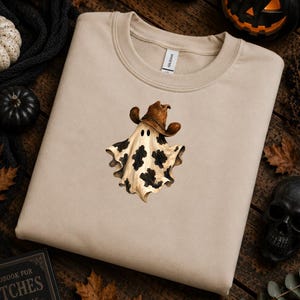 Könnte beinhalten: Ein beigefarbenes Sweatshirt mit Rundhalsausschnitt und einem zentralen Motiv eines Geistes, der einen braunen Cowboyhut trägt. Der Geist hat einen cremefarbenen Körper mit schwarzen Kuhflecken, zwei kleine schwarze Augen und wirkt fließend.