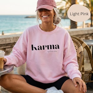 Könnte beinhalten: Hellrosa Sweatshirt mit dem Wort "Karma" in schwarzer Schrift. Darunter steht der Satz "regelt das schon" in kleinerer Schrift. Das Sweatshirt hat eine einfarbige hellrosa Farbe.