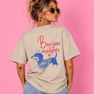 May include: A beige short-sleeved t-shirt featuring a graphic on the back. The design includes a blue and white striped dachshund wearing a beret, with the red text "Bonjour madame" and a small red heart printed above the dog illustration.