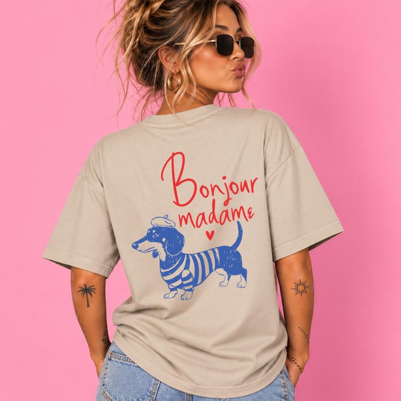 May include: A beige short-sleeved t-shirt featuring a graphic on the back. The design includes a blue and white striped dachshund wearing a beret, with the red text "Bonjour madame" and a small red heart printed above the dog illustration.