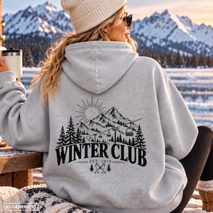 Könnte beinhalten: Ein hellgrauer Kapuzenpullover mit einem schwarzen Aufdruck auf der Rückseite. Das Motiv zeigt ein Gebirge, Tannenbäume, eine Sonne und einen Skilift. Der Text lautet WINTER CLUB EST. 1978 mit gekreuzten Skiern und kleinen Tannenbaum-Symbolen.