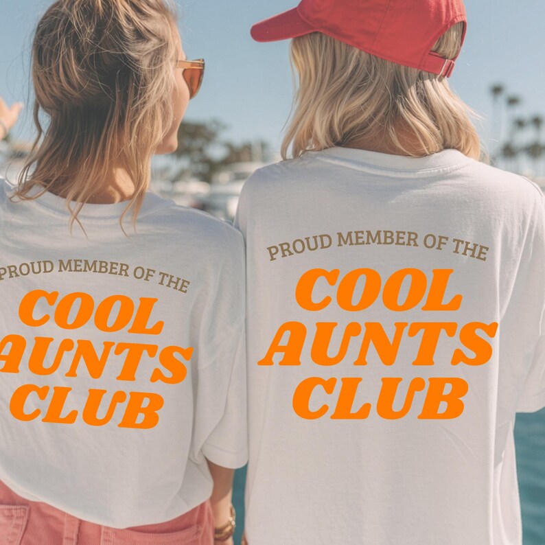 May include: Two white t-shirts with the text "PROUD MEMBER OF THE COOL AUNTS CLUB" in orange and brown lettering. The shirts are worn by two people, seen from the back, with a sunny outdoor background.