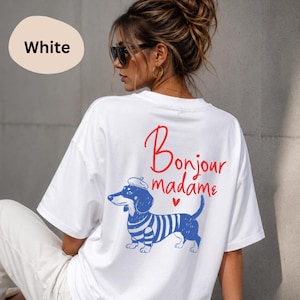 May include: A white short-sleeve t-shirt featuring a graphic on the back of a dachshund wearing a beret and a blue and white striped shirt. The text "Bonjour madame" is printed in red above the dog, accompanied by a small red heart icon.