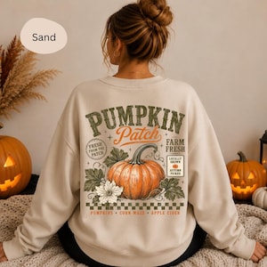 Könnte beinhalten: Ein sandfarbenes Sweatshirt mit Rundhalsausschnitt und einem großen Grafikdruck auf der Rückseite. Das Design zeigt eine zentrale orangefarbene Kürbisillustration mit grünen Blättern, umgeben von dem Text PUMPKIN Patch, FRESH FROM THE PATCH, FARM FRESH, LOCALLY GROWN, AUTUMN PICKED, PUMPKINS, CORN MAZE und APPLE CIDER.