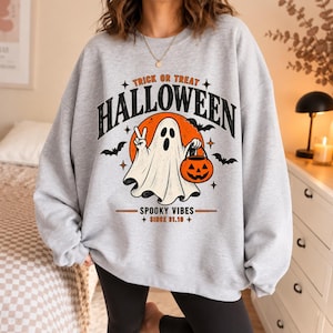 Könnte beinhalten: Ein hellgraues Sweatshirt mit Rundhalsausschnitt und einem Grafikdruck mit dem Text TRICK OR TREAT HALLOWEEN SPOOKY VIBES SINCE 31.10. Das Design zeigt ein weißes Gespenst mit einem orangefarbenen Kürbis, einen großen orangefarbenen Mond und Fledermäuse.