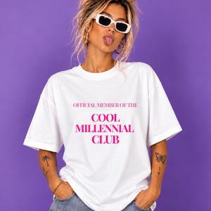 Könnte beinhalten: Ein weißes T-Shirt mit kurzen Ärmeln und dem Text "OFFICIAL MEMBER OF THE COOL MILLENNIAL CLUB", der in kräftigem Pink auf der Brust aufgedruckt ist. Das Oberteil hat einen klassischen Rundhalsausschnitt und eine lockere, bequeme Passform.