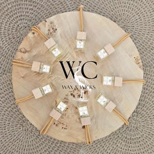 May include: A collection of small, clear glass bottles with wooden caps and tan cords, arranged on a light-colored wooden disc. The bottles are labeled and surrounded by tan cords. The disc has the text "WC Wax & Wicks" and a heron illustration.
