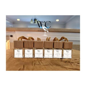 May include: Six small glass bottles with wooden caps and gold string hangers. Each bottle contains a different scented oil. The labels on the bottles read "WC", "Bare Cherry", "Brilliant Crystal", "Evergreen Apples", "Pineapple", "Plum Punch", and "Magnolia Peony".