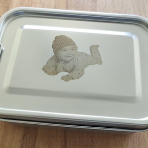 Könnte beinhalten: Eine rechteckige Lunchbox aus Edelstahl mit sicherem Schnappverschluss. Der Deckel zeigt ein eingraviertes Bild eines Babys mit gestreifter Mütze. Die Lunchbox ist für den Transport von Lebensmitteln konzipiert.