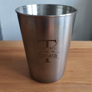 Op de afbeelding: Een roestvrijstalen beker met een gepersonaliseerde gravure. De beker heeft de naam "Tamara" onder een gestileerde letter "T" met bloemaccenten. De beker heeft een taps toelopend ontwerp en staat op een houten oppervlak.