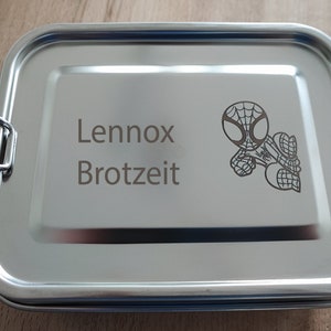 Könnte beinhalten: Eine silberne Brotdose mit einem Verschluss an jeder Seite. Die Brotdose ist mit einem eingravierten Design eines Comic-Spidermans und dem Text "Lennox Brotzeit" versehen.