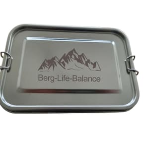 Op de afbeelding: Een rechthoekige lunchbox van roestvrij staal met een deksel. Op het deksel staat de tekst "Berg-Life-Balance" met een berg-afbeelding. De lunchbox heeft twee metalen sluitingen.