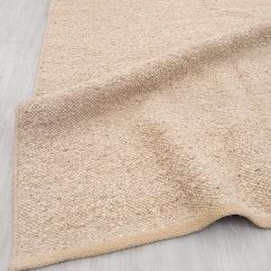Puede incluir: Una alfombra beige tejida con una superficie texturizada. La alfombra está doblada, revelando la parte inferior y el borde de la alfombra. La alfombra está hecha de una fibra natural y tiene un color neutro.
