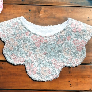May include: A white bib with a scalloped edge and a floral print in shades of pink, blue, and green. The bib has a white terry cloth lining.