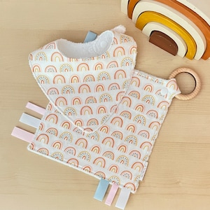May include: A white baby bib with a rainbow pattern and a wooden teether attached. The bib has a soft, absorbent fabric and is perfect for keeping baby's clothes clean during mealtime.